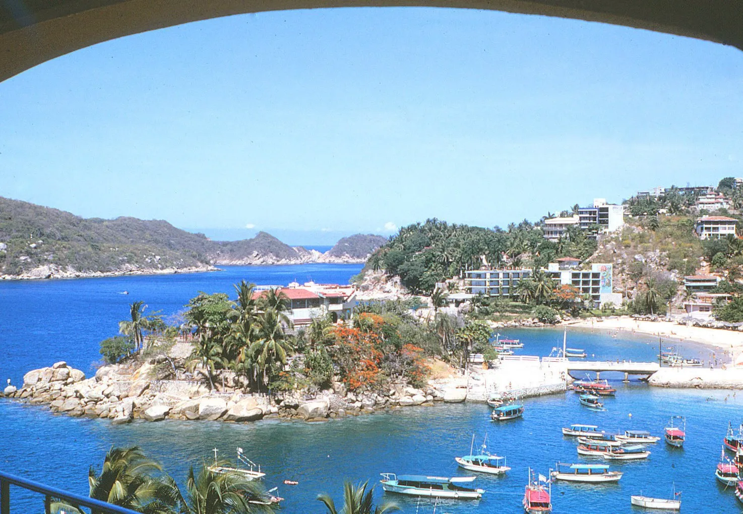 La plage de Caleta à Acapulco en 1966, vue depuis une terrasse : une baie aux eaux bleues, des barques et des collines couvertes d’hôtels