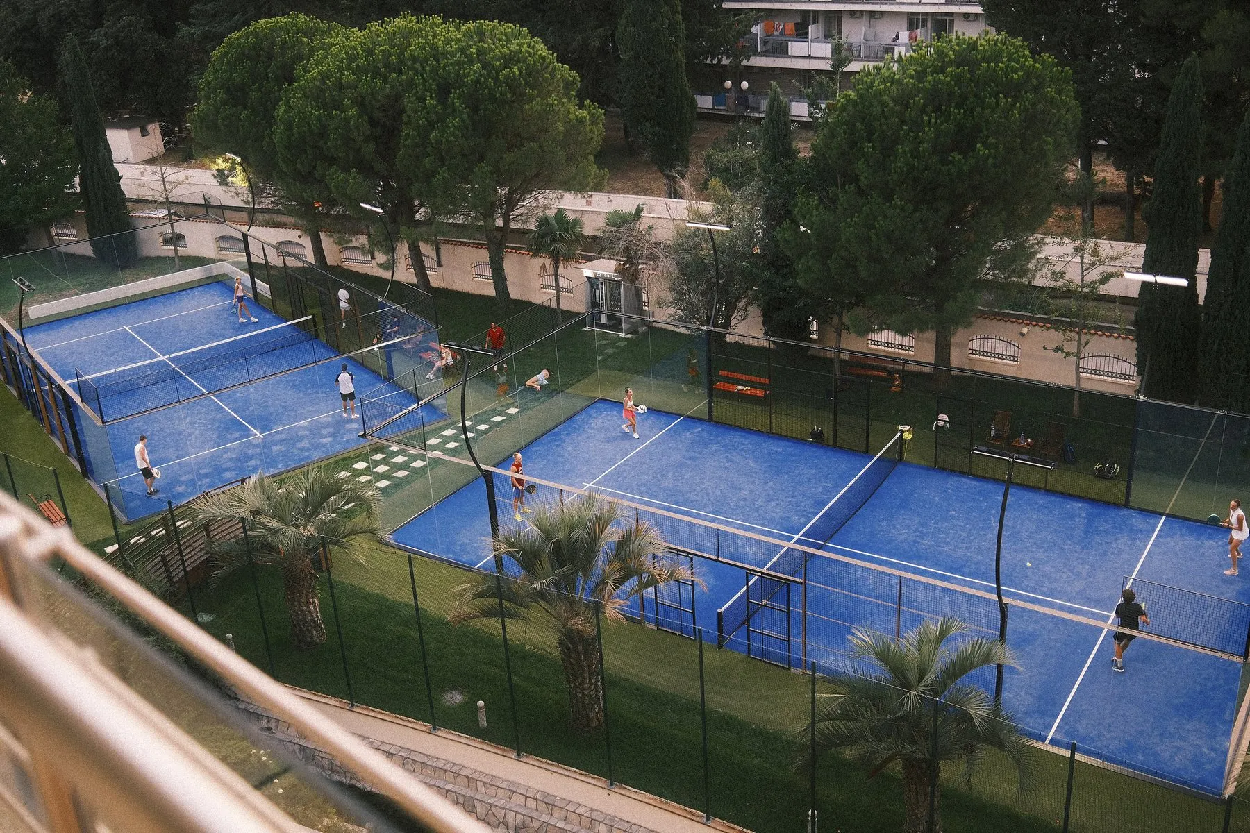 Plusieurs pistes de padel bleues entourées de verdure, avec des joueurs en double sur chacune, vues d’en haut