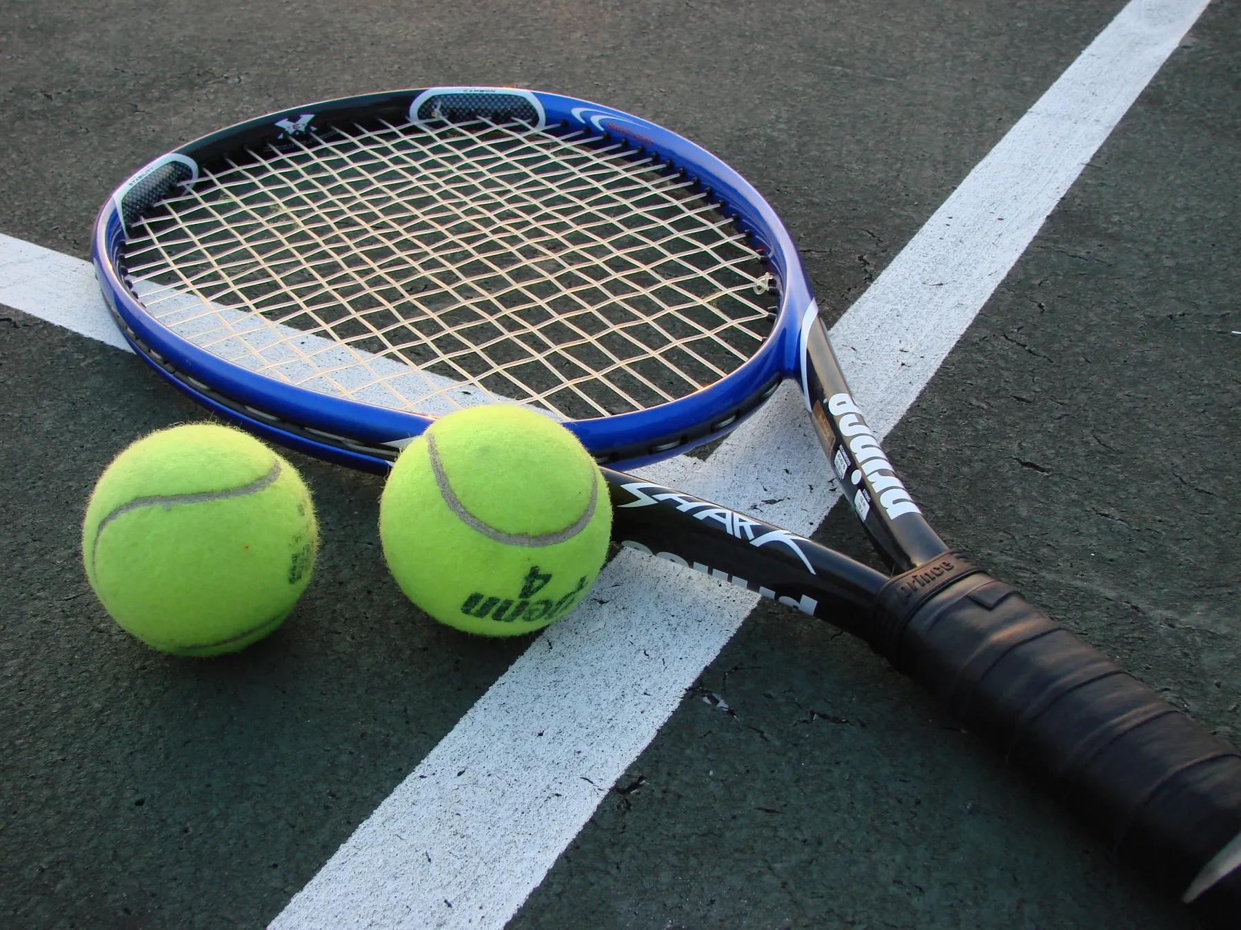 Une raquette de tennis posée sur un court en dur avec deux balles jaunes, sur une ligne blanche