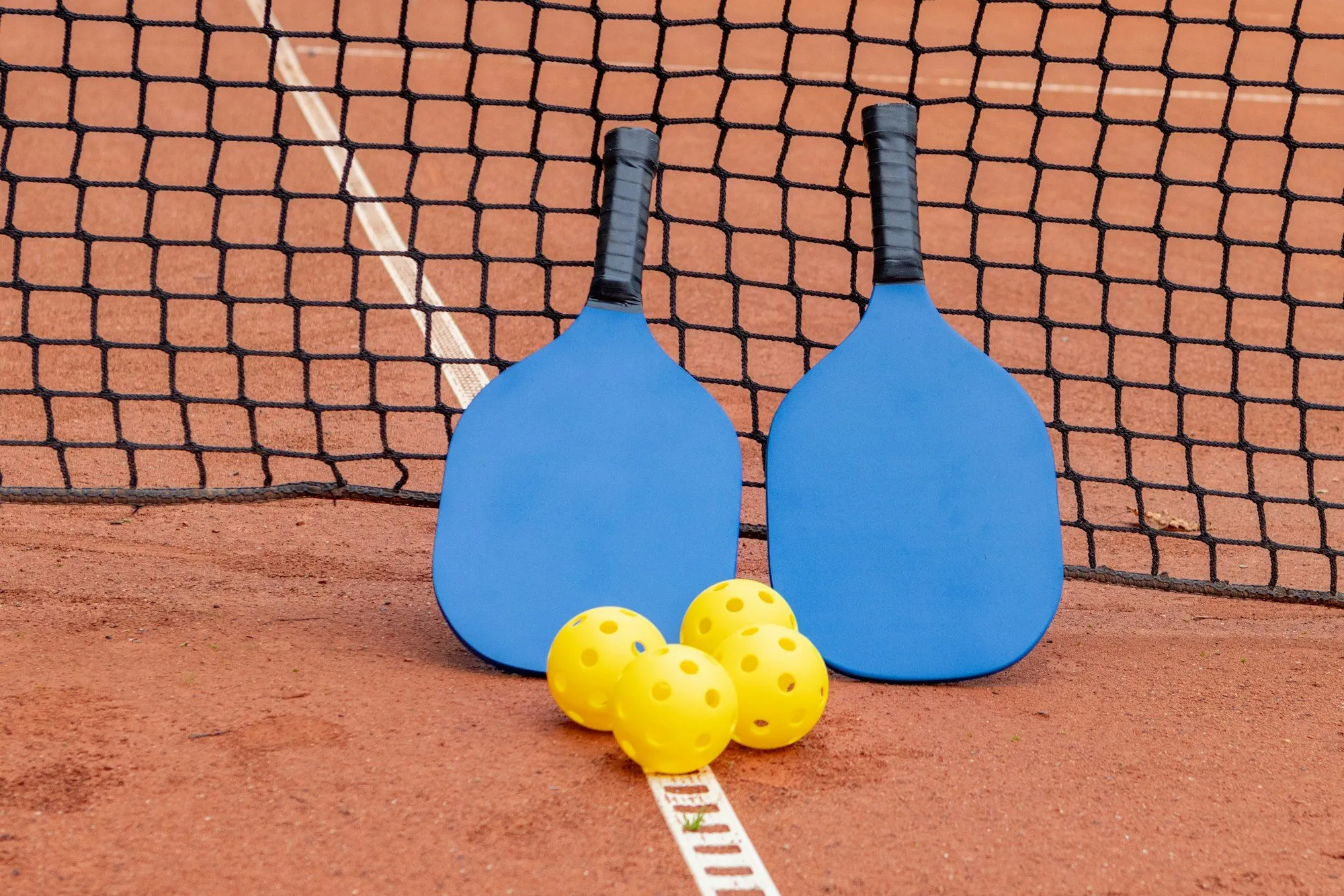 Duas raquetes de pickleball azuis e bolas amarelas perfuradas encostadas à rede de um campo de terra batida