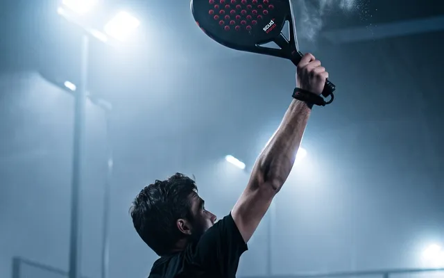 Joueur de padel au smash, raquette SCUD levée au-dessus de la tête, sur un terrain couvert éclairé de nuit