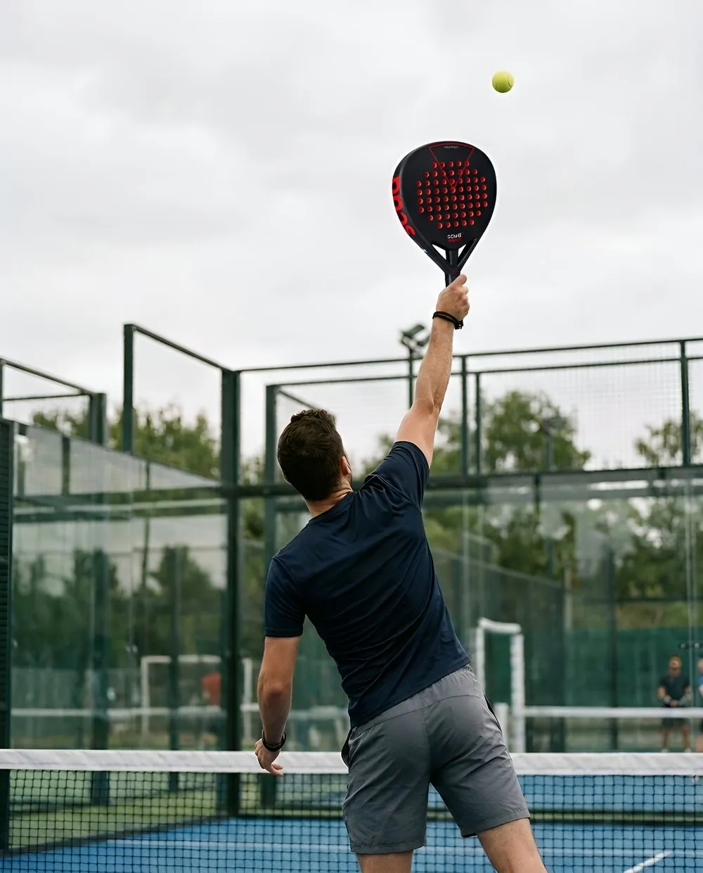Joueur de padel de dos, en tee-shirt bleu marine, bras tendu au-dessus de la tête au moment de frapper un smash avec une raquette noire à perforations rouges, sur une piste extérieure entourée de grillage