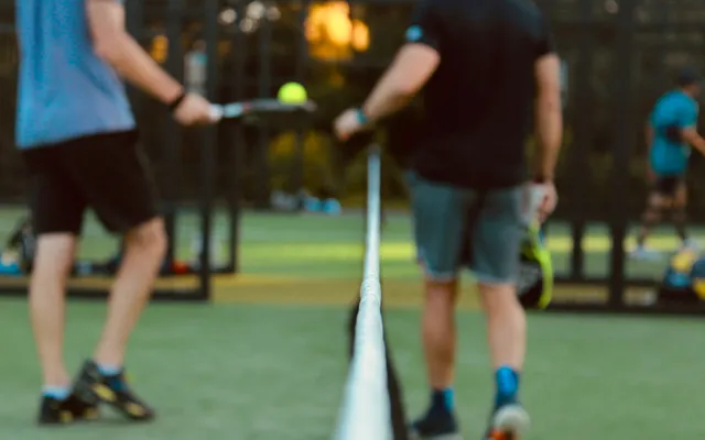 Dois jogadores de padel de cada lado da rede, bola no ar, numa luz dourada de fim de tarde