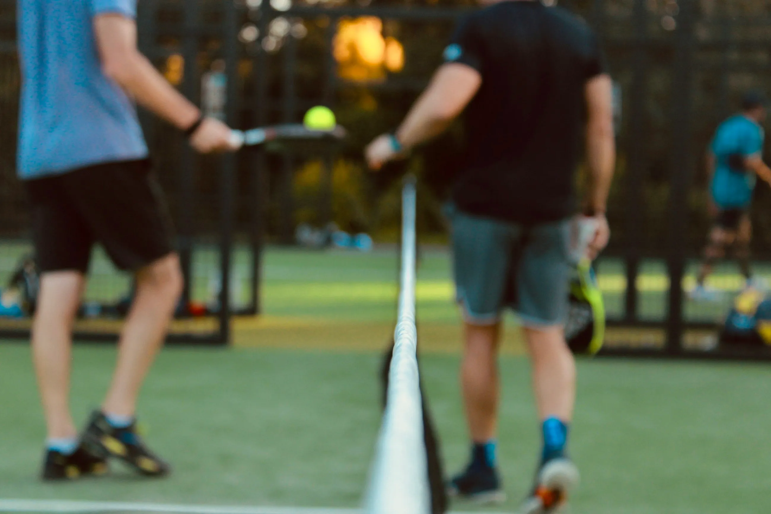 Dois jogadores de padel de cada lado da rede, bola no ar, numa luz dourada de fim de tarde