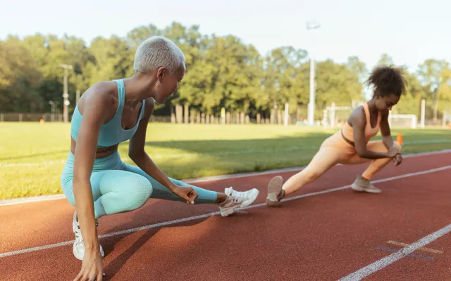 Dois atletas alongam-se em afundo lateral numa pista de atletismo ao sol da manhã