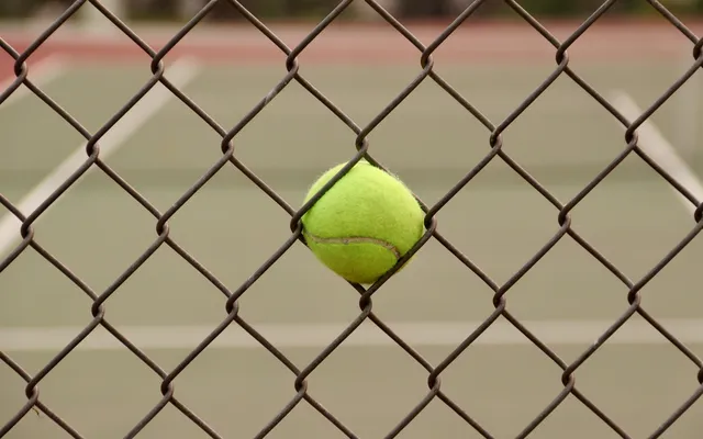 Uma bola amarela presa numa rede metálica, com o campo desfocado atrás