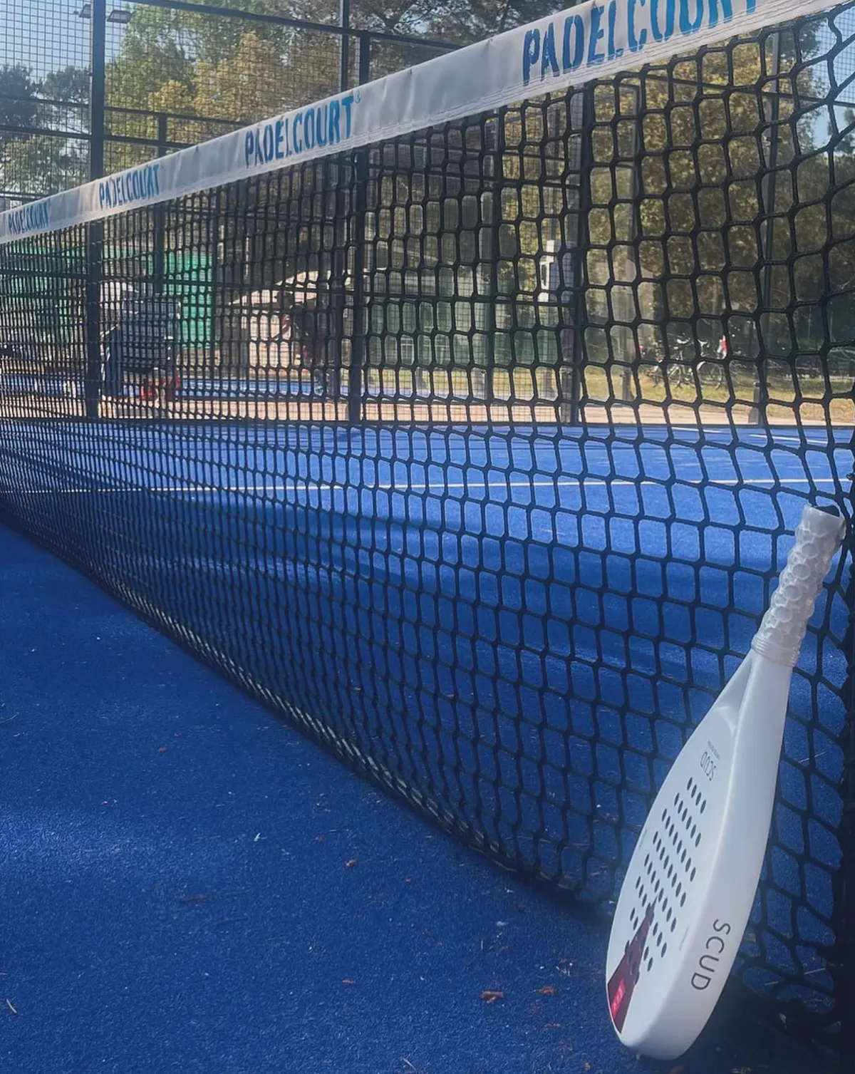 Raquette de padel SCUD blanche posée contre le filet d'un court de padel bleu, en plein soleil