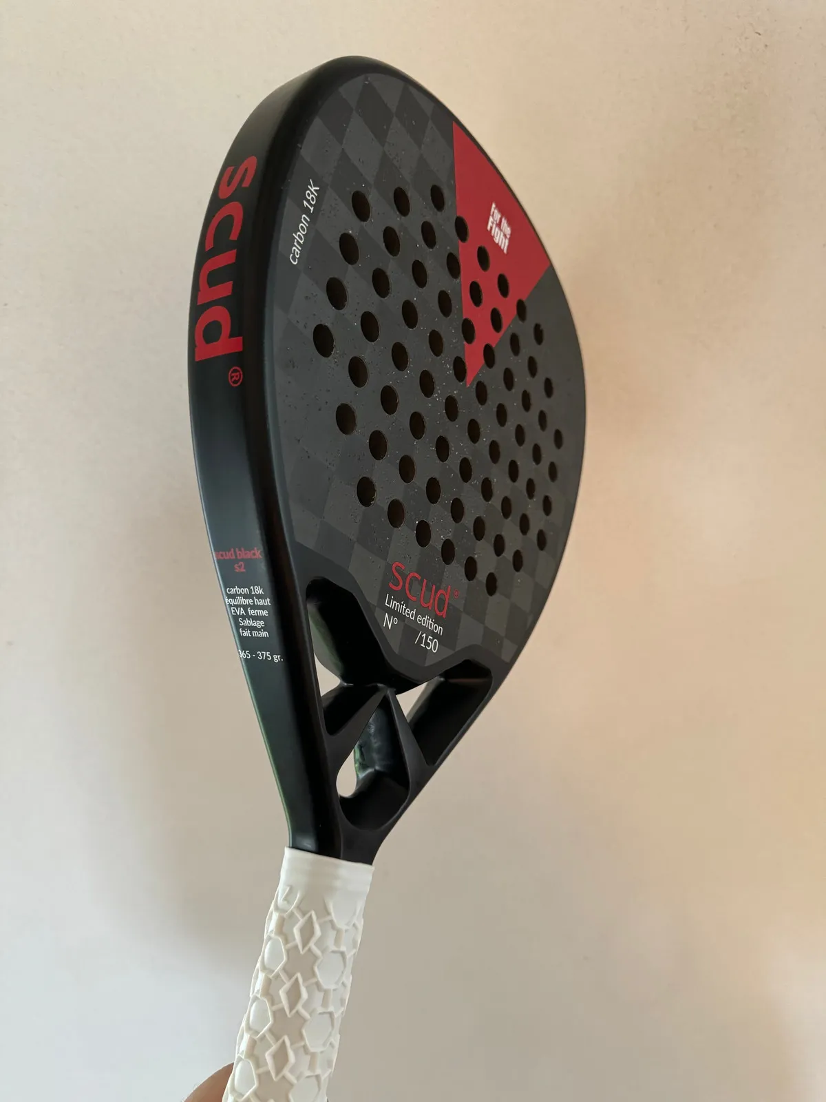 Raquette de padel SCUD noire tenue à la main devant un mur clair : le damier de carbone du tamis, le triangle rouge et le logo scud sur le cœur