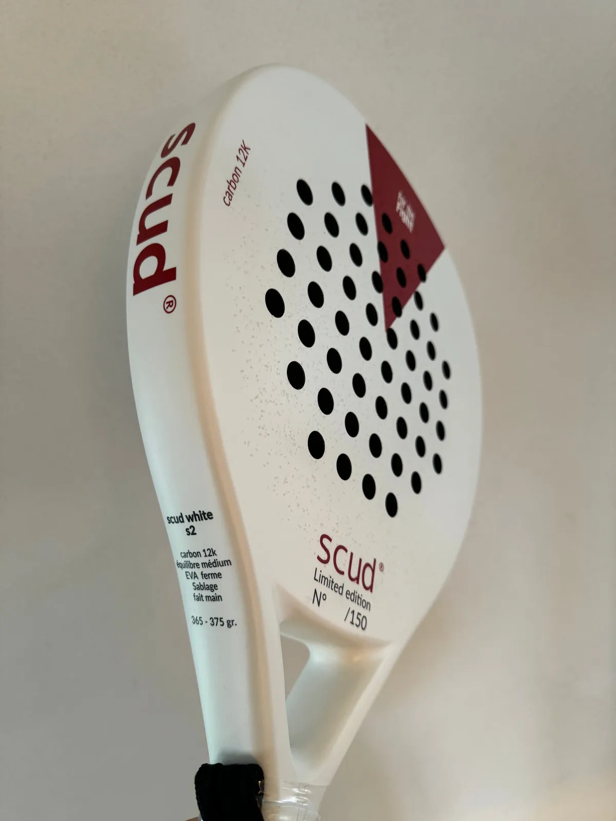 Raquette de padel SCUD blanche tenue à la main : le tamis blanc perforé, le triangle rouge et les caractéristiques imprimées le long du cadre
