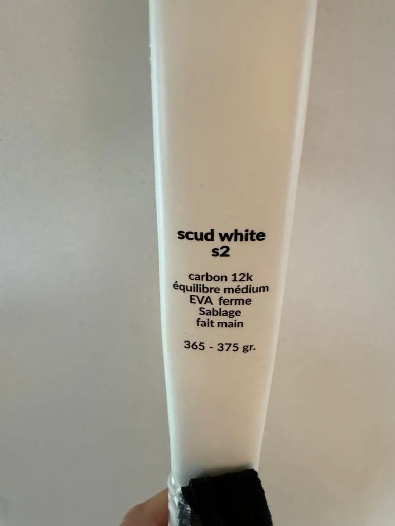 Gros plan du cadre d'une raquette de padel SCUD blanche : « scud white s2 — carbon 12k, équilibre médium, EVA ferme, sablage fait main, 365-375 gr » imprimé sous le cœur