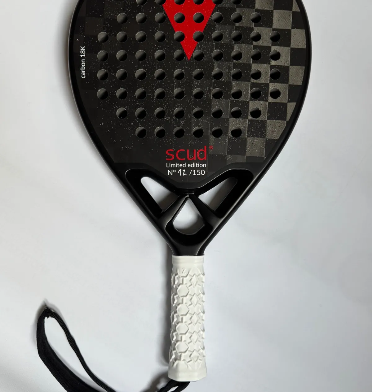 Raquette de padel SCUD noire photographiée à plat sur fond blanc : le damier de carbone 18K du tamis, le triangle rouge en tête et la mention scud Limited edition N° 12/150, grip blanc et dragonne noire