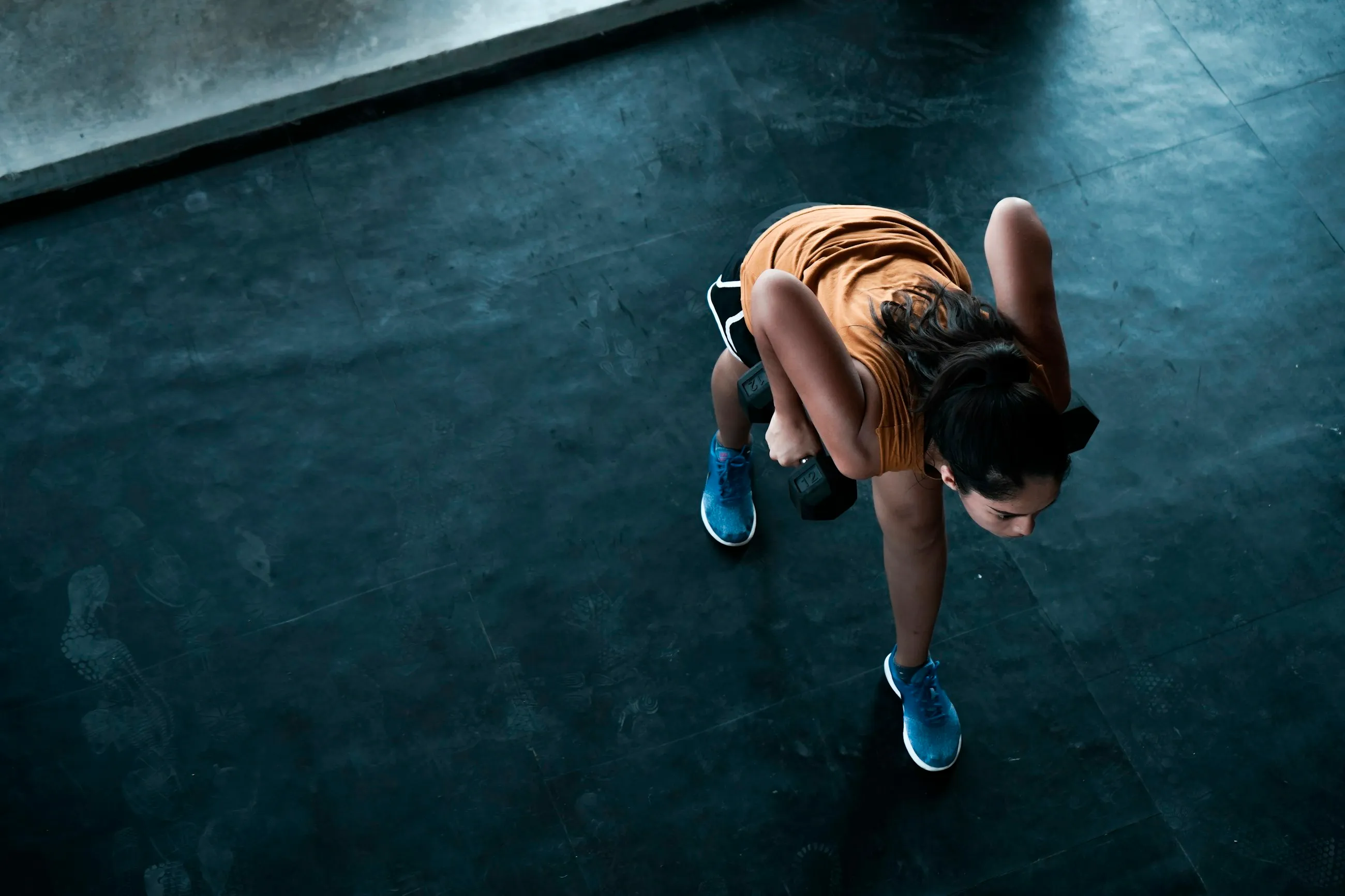 Visto de cima, um atleta em afundo atrás com dois halteres num piso preto de ginásio