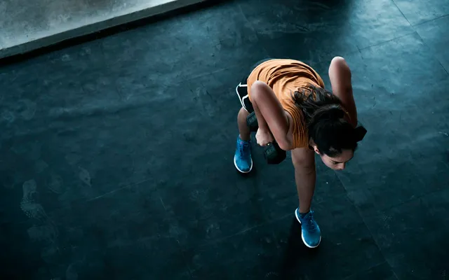Visto de cima, um atleta em afundo atrás com dois halteres num piso preto de ginásio