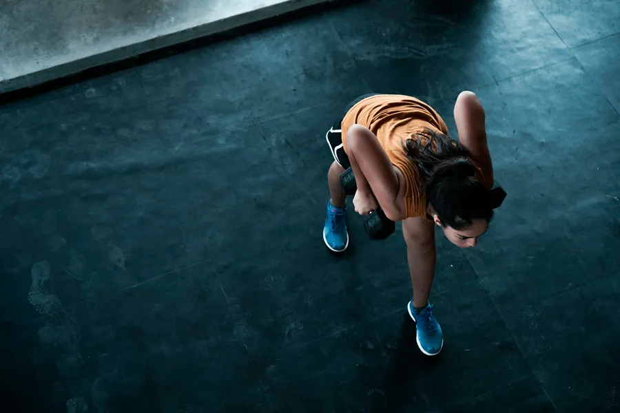 Visto de cima, um atleta em afundo atrás com dois halteres num piso preto de ginásio