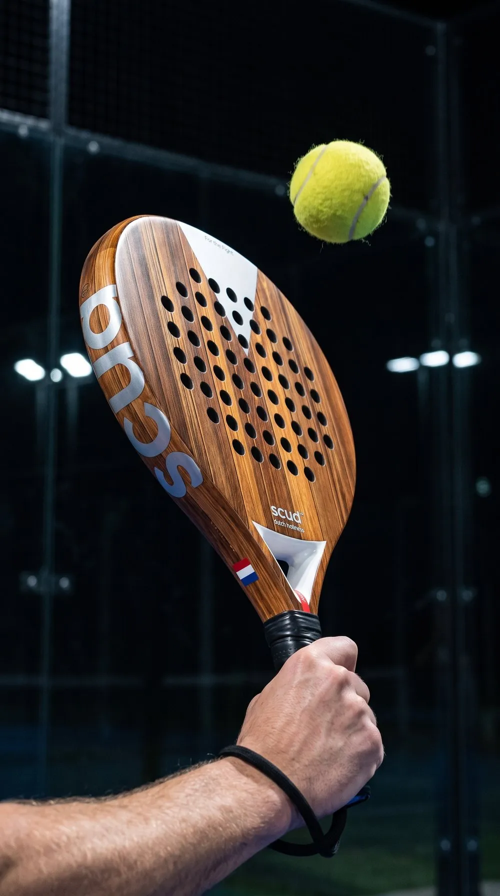 Mano que sujeta una pala SCUD con acabado de madera, la cara ligeramente inclinada, una pelota amarilla por encima y el cordón negro alrededor de la muñeca