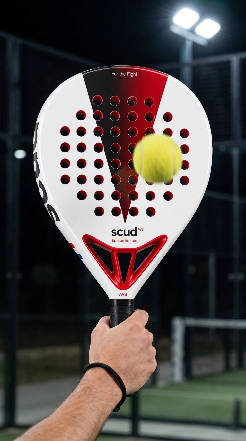 Mano que sujeta una pala SCUD blanca y roja por la parte baja del mango, la cara de frente, una pelota desenfocada delante de los agujeros y el cordón negro en la muñeca, en una pista iluminada de noche