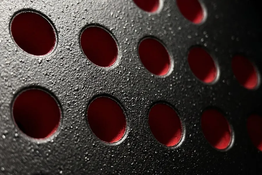 Gros plan du tamis d'une raquette de padel SCUD Black Impact B3 : perforations cerclées de rouge et cristaux de silice du sablage fait-main