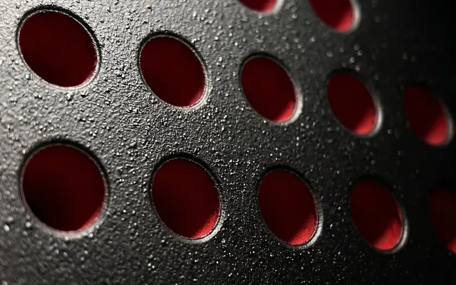 Gros plan du tamis d'une raquette de padel SCUD Black Impact B3 : perforations cerclées de rouge et cristaux de silice du sablage fait-main