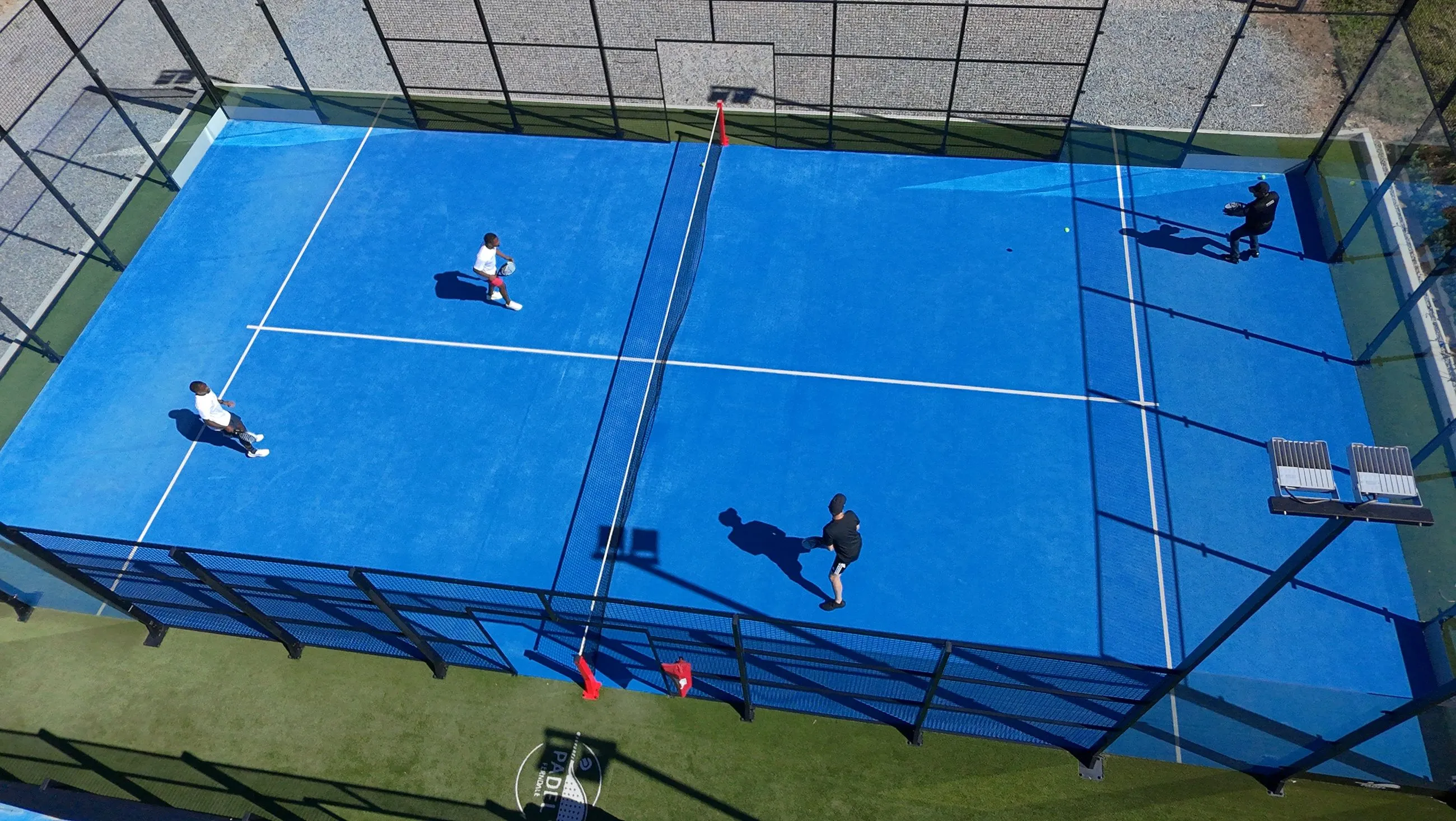 Quatro jogadores num jogo de pares num campo de padel azul ao sol, vistos de cima