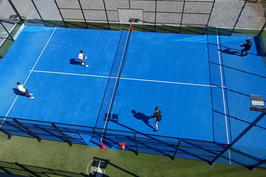Four players in a doubles match on a sunny blue padel court, seen from above